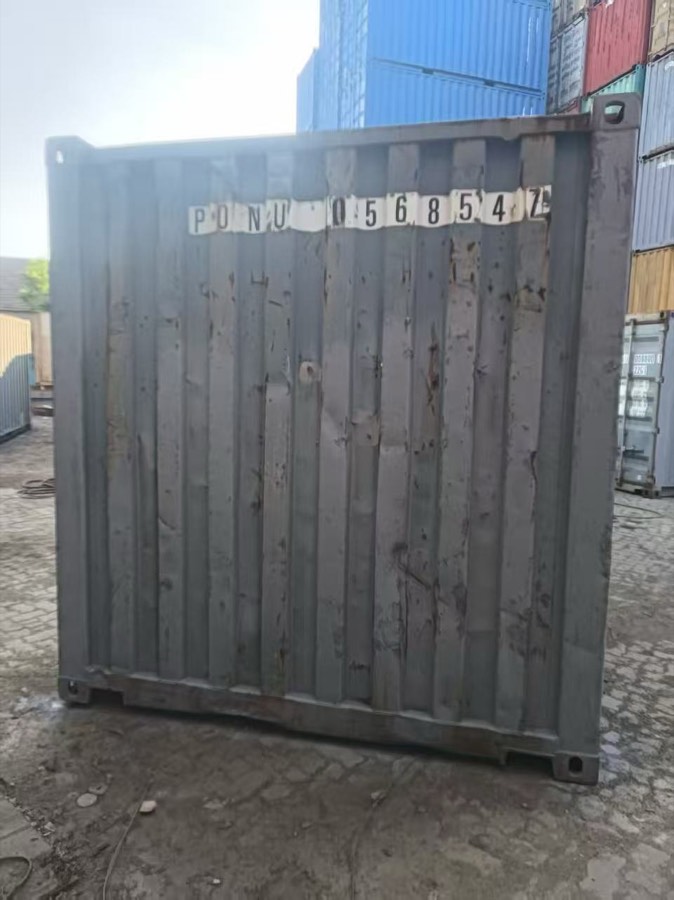 A used shipping container standing on a depot hardstand with stacked containers behind it.